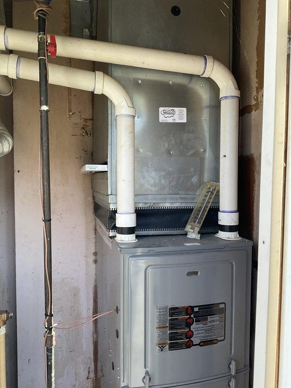 Conducted comprehensive site assessment for HVAC system replacement in residential basement utility area. Evaluated existing furnace and air handler configuration, documenting current installation including PVC venting system with multiple 90-degree bends, copper refrigerant lines, and electrical connections. Photographed complete system setup showing gray metal furnace unit with mounted air handler above, noting black coupling bands at pipe connections and red-handled valve positioning. Assessed Rheem Classic water heater in adjacent mechanical space, examining flexible ductwork connections, silver and white ribbed ducting secured with metal clamps for exhaust and intake systems. Documented copper piping connections at water heater base and reviewed overall mechanical infrastructure within exposed framing environment. Prepared detailed photographic documentation of all components for replacement estimate preparation.