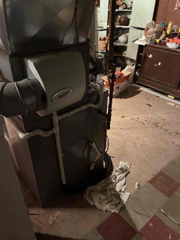 Conducted comprehensive site evaluation of existing HVAC system in basement utility area for equipment replacement estimate. Assessed current Aprilaire humidification unit with multi-component vertical configuration, connected via black flexible ductwork and copper refrigerant lines. Evaluated metallic gray furnace unit featuring lower combustion chamber with oval access panel and upper exhaust hood connection. Documented equipment specifications, ductwork routing, and mechanical infrastructure through detailed photography. Inspected surrounding utility space conditions, including concrete block walls, exposed ceiling joists with visible deterioration, and floor substrate showing wear patterns. Noted active HVAC piping, electrical connections, and ventilation pathways for replacement planning considerations. Provided customer with system replacement options and specifications for complete equipment upgrade.