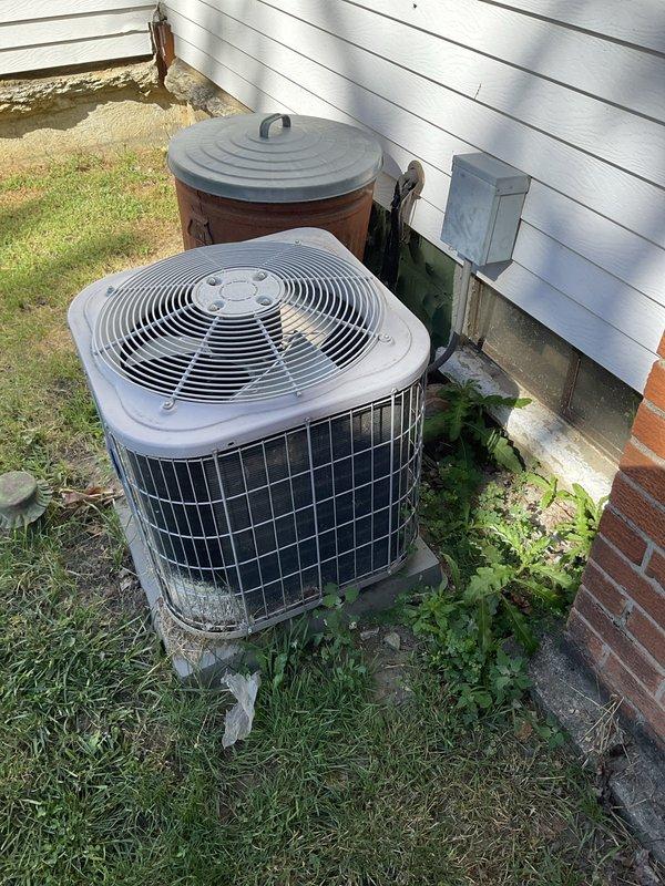Completed on-site evaluation of existing residential air conditioning system for replacement estimate. Documented current outdoor condensing unit condition, noting aging equipment with weathered components, overgrown vegetation around unit base, and installation on concrete pad adjacent to building foundation. Photographed unit placement, electrical connections, and surrounding area to assess accessibility for future replacement work and identify any potential installation considerations.