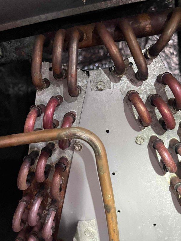 # HVAC Emergency Service Report

Responded to emergency no-cool call for 20-year-old central air conditioning system blowing warm air. Upon arrival, performed diagnostic assessment of the outdoor condensing unit and indoor evaporator coil assembly. 

Inspection revealed severely aged copper refrigerant lines with extensive oxidation, corrosion, and patina buildup throughout the refrigerant circuit. Multiple refrigerant lines passing through the evaporator coil mounting plate showed varying degrees of deterioration, with discoloration ranging from bright copper to dark brown indicating prolonged exposure to heat and moisture cycling. The distribution plate connections displayed significant tarnishing and corrosion consistent with a system operating beyond its expected service life.

Visual examination of the confined mechanical space showed heavy dust accumulation and environmental degradation of copper tubing. The varying oxidation levels across different pipe sections suggested possib