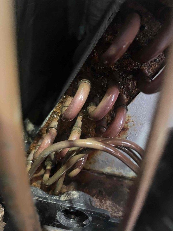 # HVAC Emergency Service Report

Responded to emergency no-cool call for 20-year-old central air conditioning system blowing warm air. Upon arrival, performed diagnostic assessment of the outdoor condensing unit and indoor evaporator coil assembly. 

Inspection revealed severely aged copper refrigerant lines with extensive oxidation, corrosion, and patina buildup throughout the refrigerant circuit. Multiple refrigerant lines passing through the evaporator coil mounting plate showed varying degrees of deterioration, with discoloration ranging from bright copper to dark brown indicating prolonged exposure to heat and moisture cycling. The distribution plate connections displayed significant tarnishing and corrosion consistent with a system operating beyond its expected service life.

Visual examination of the confined mechanical space showed heavy dust accumulation and environmental degradation of copper tubing. The varying oxidation levels across different pipe sections suggested possib