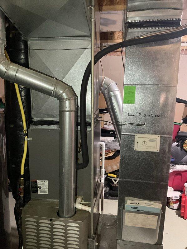 Completed full HVAC system replacement installation in basement utility area, including new Carrier furnace and air handler unit positioned side by side. Installed metallic ductwork with 90-degree elbow joint and black insulation wrapping, connected flexible ducting to air handler, and mounted wall thermostat on concrete block wall for system control. All components properly secured, labeled, and tested for operational readiness.