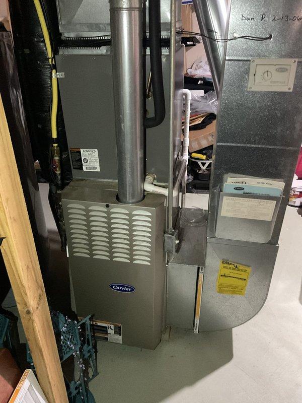 Completed full HVAC system replacement installation in basement utility area, including new Carrier furnace and air handler unit positioned side by side. Installed metallic ductwork with 90-degree elbow joint and black insulation wrapping, connected flexible ducting to air handler, and mounted wall thermostat on concrete block wall for system control. All components properly secured, labeled, and tested for operational readiness.