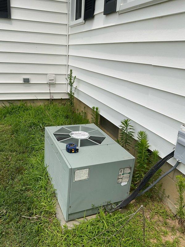 Conducted on-site evaluation of existing outdoor air conditioning condenser unit for replacement estimate. Inspected light green/sage colored AC unit positioned on ground-level pad adjacent to house foundation, documenting current system configuration including refrigerant line routing, electrical connections, and unit placement. Photographed equipment from multiple angles to capture unit condition, surrounding clearances, ventilation access, and existing line set installation for accurate replacement system sizing and planning purposes.