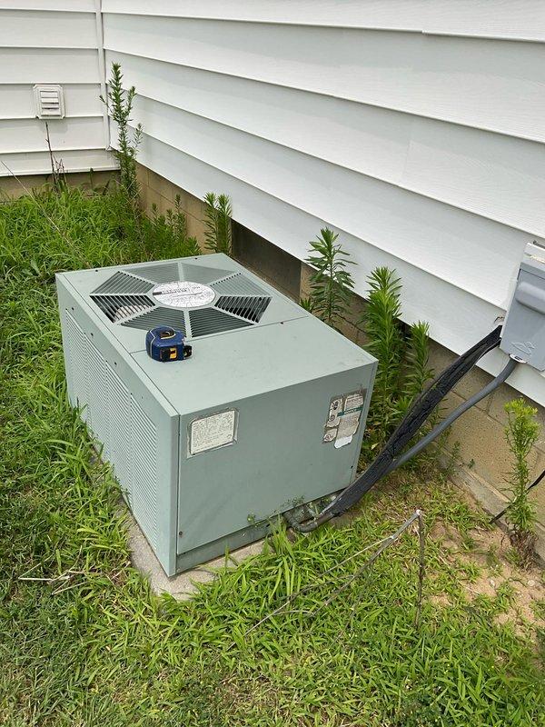 Conducted on-site evaluation of existing outdoor air conditioning condenser unit for replacement estimate. Inspected light green/sage colored AC unit positioned on ground-level pad adjacent to house foundation, documenting current system configuration including refrigerant line routing, electrical connections, and unit placement. Photographed equipment from multiple angles to capture unit condition, surrounding clearances, ventilation access, and existing line set installation for accurate replacement system sizing and planning purposes.