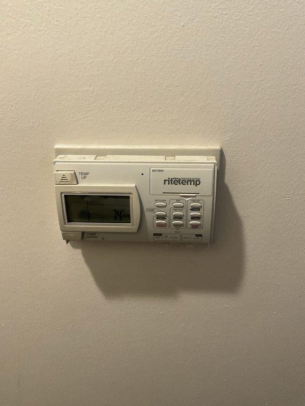 Performed comprehensive air conditioning replacement estimate for aging residential cooling system. Evaluated existing outdoor condensing unit showing significant deterioration with visible rust and corrosion on disconnect box, metal conduits, and mounting components. Unit confirmed to be approximately fifteen years old, exceeding typical service life expectancy. Inspected indoor programmable Ritetemp thermostat for compatibility assessment with potential replacement equipment. Documented current system configuration including outdoor unit placement on gravel pad with metal ring border adjacent to property structure, electrical disconnect condition, and refrigerant line routing. Photographed equipment conditions to support replacement recommendations. Customer requesting complete system replacement proposal.