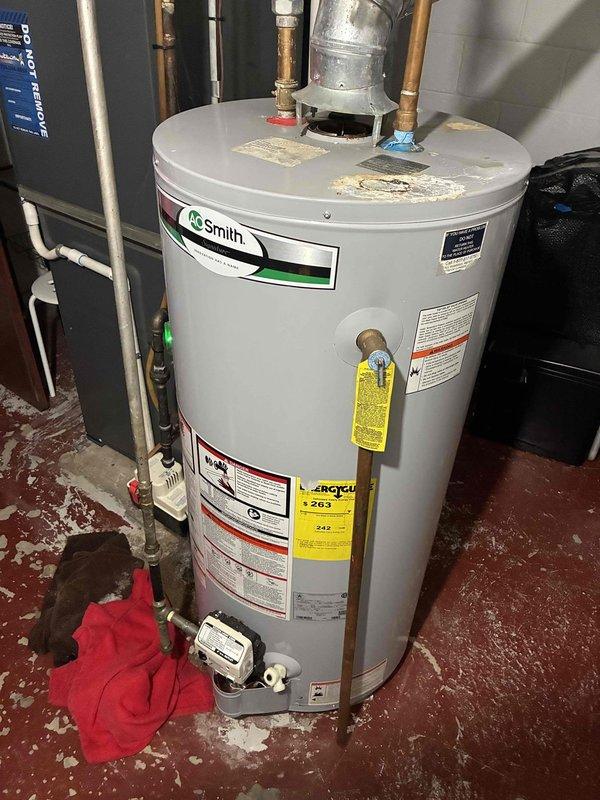 Conducted on-site assessment of non-functioning A.O. Smith standard natural draft water heater located in basement utility area. Evaluated existing unit configuration including copper supply piping, flue venting system, pressure relief valve assembly, and electrical connections. Documented current installation conditions and measured available space for replacement unit, noting concrete block wall construction and existing plumbing layout with PVC and copper pipe runs.