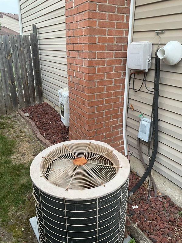 Conducted on-site evaluation of existing cylindrical air conditioning condenser unit for replacement estimate with both homeowners present. Assessed aging outdoor unit showing significant wear, surface discoloration on housing panels, and visible rust/corrosion on central fan hub components. Documented current installation configuration including concrete pad placement, electrical disconnect box positioning, refrigerant line routing along vinyl siding, and clearance requirements adjacent to brick chimney and wooden privacy fence. Photographed unit condition from multiple angles to capture weathering patterns, black horizontal slat deterioration, and surrounding landscape elements including mulch bed and grass areas. Evaluated electrical conduit connections and discussed replacement options for complete system upgrade.