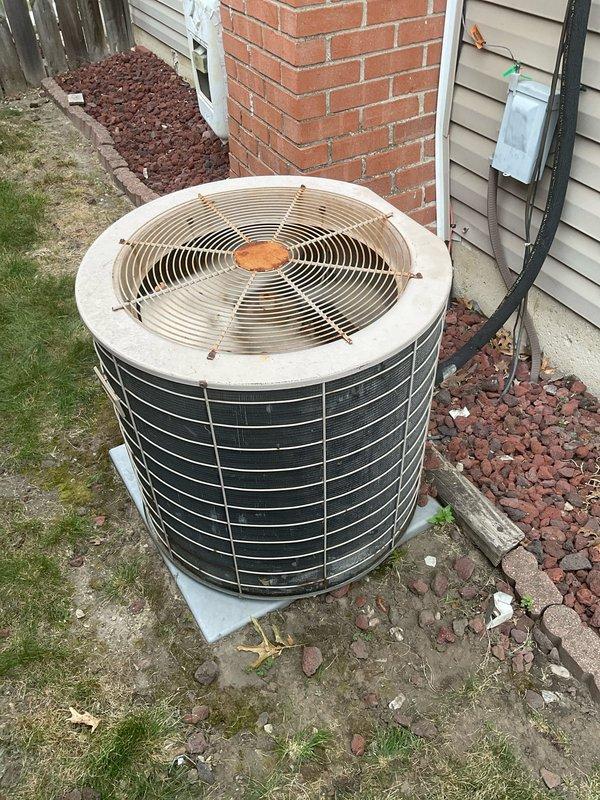 Conducted on-site evaluation of existing cylindrical air conditioning condenser unit for replacement estimate with both homeowners present. Assessed aging outdoor unit showing significant wear, surface discoloration on housing panels, and visible rust/corrosion on central fan hub components. Documented current installation configuration including concrete pad placement, electrical disconnect box positioning, refrigerant line routing along vinyl siding, and clearance requirements adjacent to brick chimney and wooden privacy fence. Photographed unit condition from multiple angles to capture weathering patterns, black horizontal slat deterioration, and surrounding landscape elements including mulch bed and grass areas. Evaluated electrical conduit connections and discussed replacement options for complete system upgrade.