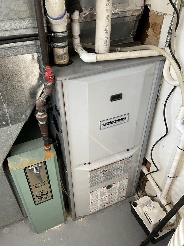 Conducted on-site assessment of existing residential HVAC system for replacement estimate. Inspected Comfortmaker furnace/air handler unit in basement utility area, evaluating overall condition of equipment, ductwork, and associated components. Documented aging galvanized metal ductwork showing dust accumulation and discoloration, existing white cabinet furnace with digital control panel, and auxiliary equipment including light blue-green condensate basin. Examined plumbing connections including PVC vent piping, copper lines, and shut-off valves for integration with new equipment installation. Photographed current system configuration, noting concrete block wall damage near equipment area that may require attention during replacement. Provided comprehensive equipment replacement recommendations based on existing infrastructure and spatial constraints.