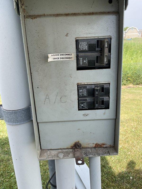 Conducted on-site evaluation for air conditioning system replacement. Assessed existing outdoor condensing unit with cylindrical design and wire mesh grille showing significant weathering, discoloration, and age-related deterioration. Inspected electrical disconnect box mounted on exterior wall, noting corroded condition with rust spots and surface degradation. Documented current equipment configuration including PVC conduit routing and concrete pad placement adjacent to structure. Photographed existing system components to provide comprehensive replacement estimate for new AC installation.
