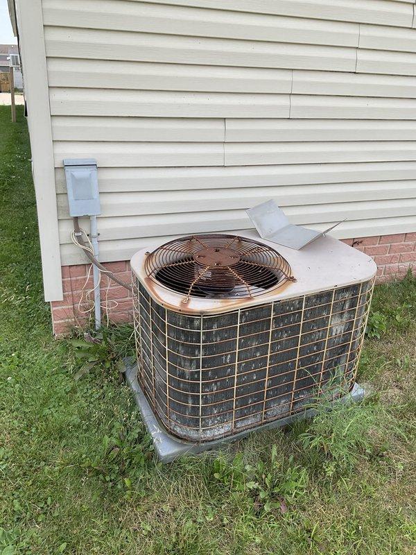 Conducted on-site evaluation for air conditioning system replacement. Assessed existing outdoor condensing unit with cylindrical design and wire mesh grille showing significant weathering, discoloration, and age-related deterioration. Inspected electrical disconnect box mounted on exterior wall, noting corroded condition with rust spots and surface degradation. Documented current equipment configuration including PVC conduit routing and concrete pad placement adjacent to structure. Photographed existing system components to provide comprehensive replacement estimate for new AC installation.