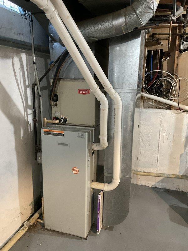 Conducted on-site evaluation of 20-year-old R22 air conditioning system that was no longer adequately cooling the residence. Documented existing Bryant furnace installation in basement utility area along with associated ductwork, condensate drainage lines, and electrical connections. Photographed outdoor condensing unit positioned on concrete pad with electrical meter and service connections. System assessment confirmed age-related performance decline and obsolete refrigerant type. Prepared replacement estimate for basic air conditioning system installation suitable for property transfer to future occupants.