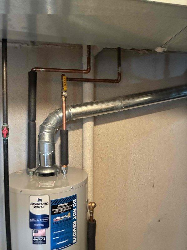 Completed installation of Bradford White water heater in basement utility area. Unit properly positioned and secured with all copper supply lines connected, pressure relief valve installed, and metallic flue vent pipe routed through ceiling with appropriate elbows and support brackets. All connections tested and insulation applied to water lines to prevent condensation and heat loss.