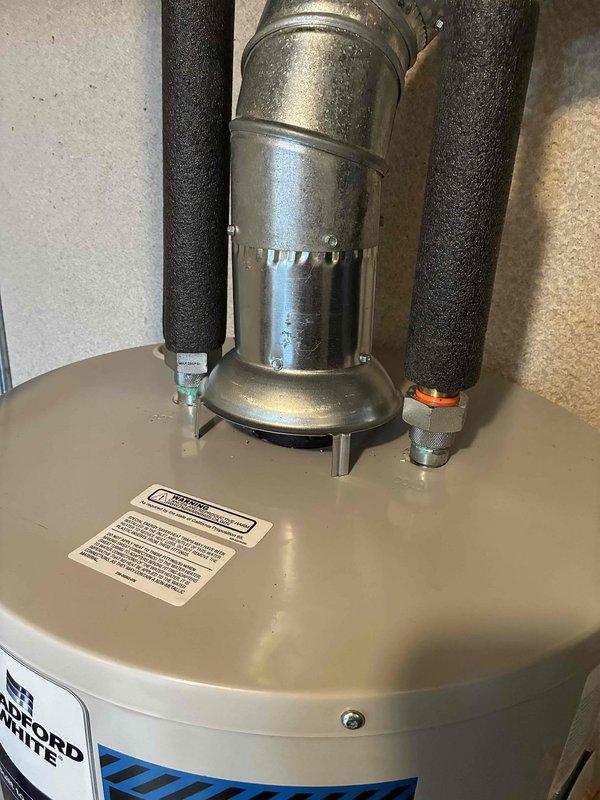 Completed installation of Bradford White water heater in basement utility area. Unit properly positioned and secured with all copper supply lines connected, pressure relief valve installed, and metallic flue vent pipe routed through ceiling with appropriate elbows and support brackets. All connections tested and insulation applied to water lines to prevent condensation and heat loss.