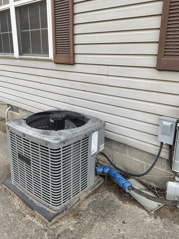 Performed on-site assessment of existing residential air conditioning system for mobile home office facility at campground location. Evaluated outdoor condensing unit condition, including inspection of cabinet integrity, fan assembly, and refrigerant line insulation. Documented current equipment specifications, electrical disconnect configuration, and mounting platform condition. Photographed installation setup showing unit positioning relative to structure, utility connections including electrical meter and panel box, and PVC condensate drainage routing. Noted blue foam pipe insulation sections present near unit base and assessed overall system accessibility for potential replacement work. Provided comprehensive equipment evaluation and replacement estimate for single-component AC system upgrade.