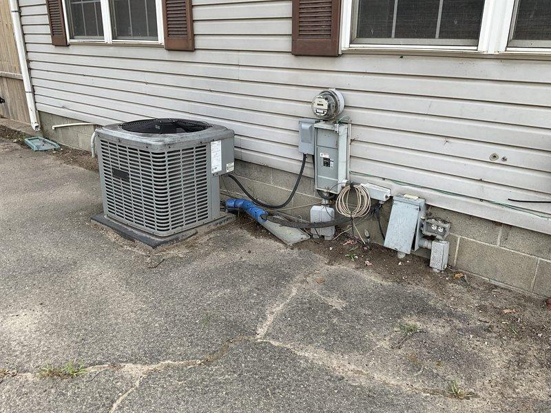 Performed on-site assessment of existing residential air conditioning system for mobile home office facility at campground location. Evaluated outdoor condensing unit condition, including inspection of cabinet integrity, fan assembly, and refrigerant line insulation. Documented current equipment specifications, electrical disconnect configuration, and mounting platform condition. Photographed installation setup showing unit positioning relative to structure, utility connections including electrical meter and panel box, and PVC condensate drainage routing. Noted blue foam pipe insulation sections present near unit base and assessed overall system accessibility for potential replacement work. Provided comprehensive equipment evaluation and replacement estimate for single-component AC system upgrade.
