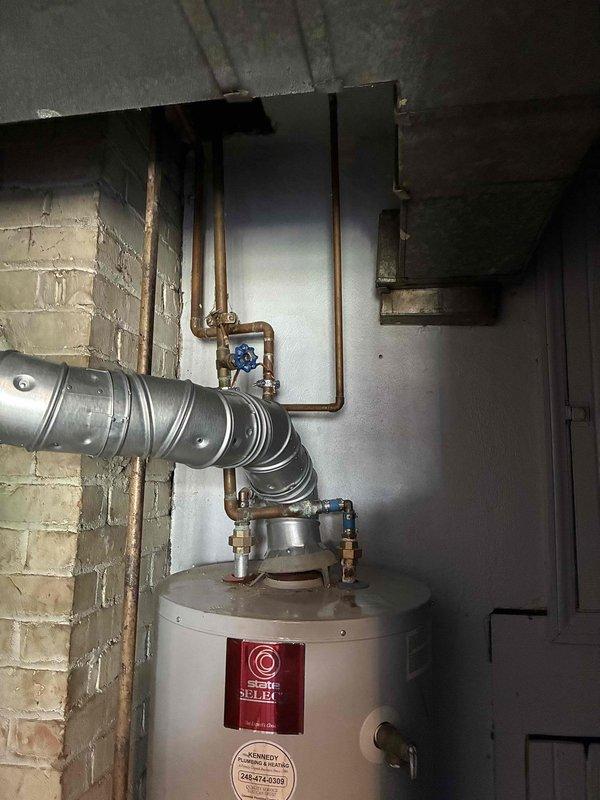 Responded to emergency service call for non-functional tanked water heater replacement in residential basement utility room. Assessed existing white cylindrical water heater unit with power vent configuration installed against painted cinder block wall on concrete floor. Provided comprehensive estimate for complete water heater replacement including removal of failed unit, installation of new tanked water heater with proper venting through existing flexible metallic duct system, connection to existing copper supply lines with blue-handled shutoff valves, and compliance with current plumbing codes for basement installations.