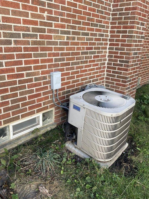 Conducted comprehensive site assessment for complete HVAC system replacement at residential property. Evaluated existing outdoor condensing unit positioned on concrete pad against brick exterior, noting cylindrical configuration with vertical cooling fins and top-mounted fan assembly. Inspected electrical disconnect box and conduit connections serving outdoor equipment. Assessed indoor basement infrastructure including electrical panel, existing ductwork, and utility area layout to determine installation requirements for new furnace and air handler. Documented current system configuration and measured available space for equipment replacement. Photographed existing equipment conditions and electrical service to support replacement proposal specifications.