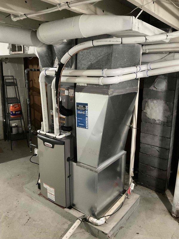 Completed full HVAC system installation in basement mechanical room, including 70,000 BTU forced-air furnace with beige metal cabinet, 1.5-ton air conditioning unit with rear discharge configuration, and whole-home humidifier. Installed gray metal air handler with proper plenum connections and large rectangular supply ductwork wrapped in silver insulation. Routed white PVC condensate and vent piping overhead with appropriate black hangers and support straps secured to ceiling joists. Mounted equipment on concrete pad at floor level with proper clearances. Connected all ductwork branches and performed electrical reconnection. System tested and verified operational upon completion.