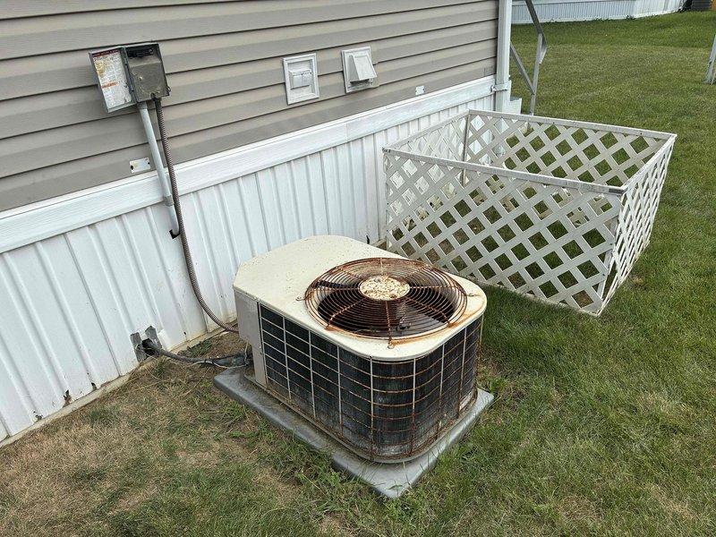 Conducted on-site evaluation of existing HVAC system at mobile home residence to provide air conditioning replacement estimate. Inspected outdoor condenser unit showing signs of age and weathering, with visible dirt accumulation and surface discoloration on beige cabinet and black louvered panels. Assessed unit positioning on concrete pad against exterior vinyl siding with lattice screening. Evaluated interior mechanical thermostat for system compatibility considerations. Documented equipment condition and installation environment through photographs for accurate proposal development.