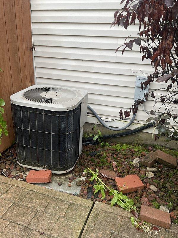 Conducted on-site evaluation of existing outdoor air conditioning unit with cylindrical condenser configuration and white top panel. Assessed current system condition, documented equipment specifications, and photographed installation for replacement estimate preparation. Unit located on concrete paver patio against vinyl-sided residence with PVC refrigerant line set routing along exterior wall.