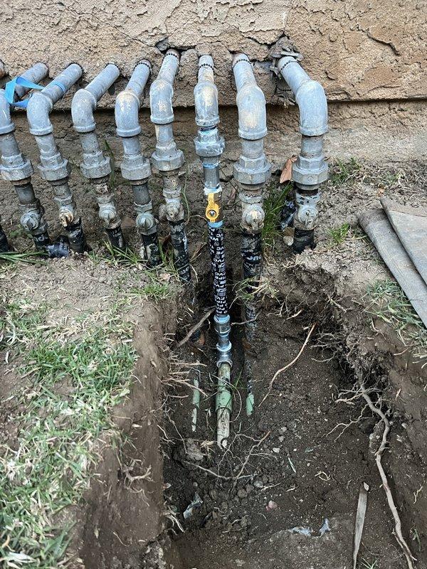 Excavated trench to access and inspect the exterior plumbing system consisting of multiple PVC supply lines with associated valves. Documented condition of the aging distribution system, noting visible weathering, mineral deposits and minor corrosion on metal valve components where the main line branches to multiple distribution points.