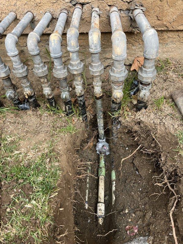 Excavated trench to access and inspect the exterior plumbing system consisting of multiple PVC supply lines with associated valves. Documented condition of the aging distribution system, noting visible weathering, mineral deposits and minor corrosion on metal valve components where the main line branches to multiple distribution points.