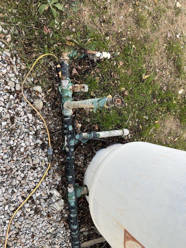 Inspected outdoor plumbing installation consisting of a large white cylindrical tank with connected black insulated pipes and copper manifold system. Documented significant oxidation and verdigris formation on copper components. Photographed installation to record current condition of valves, fittings and pipe insulation for assessment of weathering and potential maintenance needs.