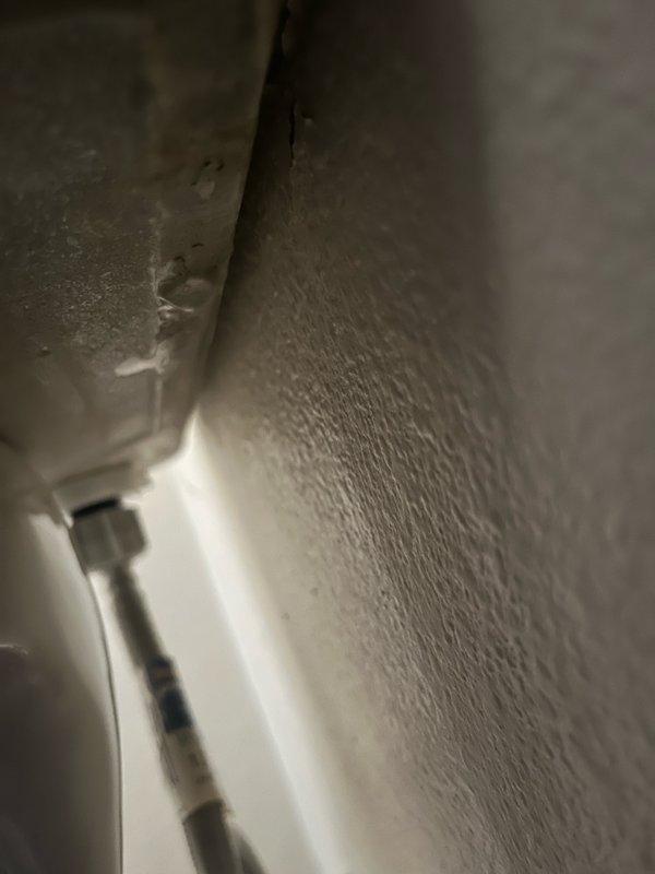 Inspected wall junction and plumbing fixture showing signs of moisture damage. Documented condition of float valve assembly with red cap and adjacent drain pipe. Observed possible mineral deposits or early mold formation on textured surface near pipe conduit.