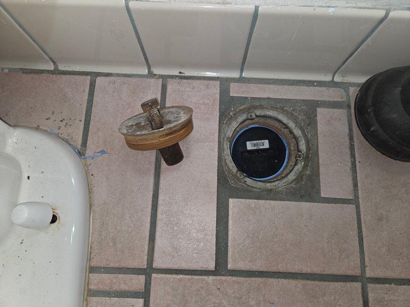 Performed inspection and maintenance of bathroom floor drain. Removed drain cover to access interior components, revealing significant corrosion and mineral buildup around the drain assembly. Identified a blue pipe fitting within the drain housing that required attention. Documentation photos captured the condition of the drain system and surrounding floor area for service records.