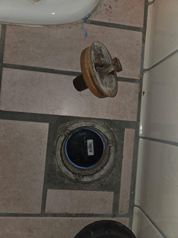 Performed inspection and maintenance of bathroom floor drain. Removed drain cover to access interior components, revealing significant corrosion and mineral buildup around the drain assembly. Identified a blue pipe fitting within the drain housing that required attention. Documentation photos captured the condition of the drain system and surrounding floor area for service records.