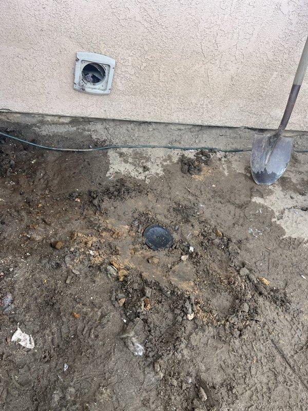 Excavated soil around exterior wall junction to access utility connection point. Exposed and inspected cable/wire running along foundation, documenting condition of weathered stucco wall with visible cracking. Located and partially uncovered round access cover embedded in ground for service connection.