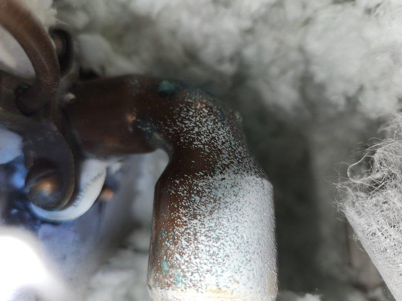 Responded to report of suspected main water line leak following notification of high water usage from municipal utility. Conducted comprehensive inspection of water main system at residential property. Located and examined main water line, identifying significant corrosion and mineral deposit accumulation on metallic piping components. Observed extensive white crystalline buildup and surface deterioration on curved pipe section, with visible blue-green oxidation patterns indicating advanced corrosion. Pipe exhibited moisture presence and compromised structural integrity consistent with active leak conditions. Documented findings with photographic evidence showing deteriorated pipe condition within insulated wall cavity or crawlspace area. Confirmed leak source as corroded section of approximately 20-year-old main water line. Provided homeowner with diagnostic findings and recommended repair options for compromised water main section.