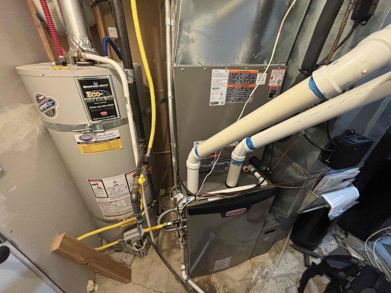 Completed Gold Cooling Maintenance on a 9-month-old residential HVAC system with equipment located in the basement and outside at the side of the house. Performed comprehensive system inspection including examination of furnace components, gas valve assembly, electrical connections, blower motor housing, and condensate drain lines, verifying all connections were intact and properly secured. Documented equipment condition with photos showing proper installation and maintenance of water heater, furnace unit, and associated PVC piping and utility lines.