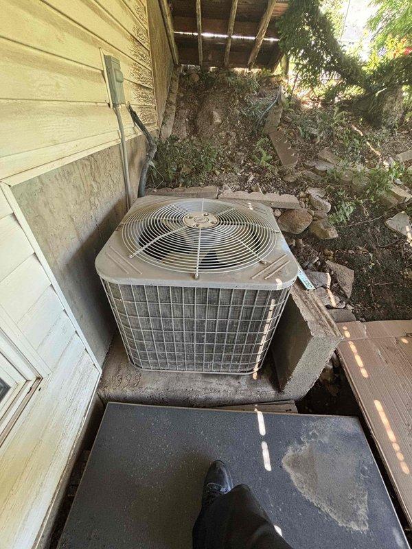 Responded to no cooling complaint on residential single-zone air conditioning system over ten years old with condenser unit located in backyard. Upon arrival, found unit producing warm air at supply vents and condenser making abnormal noises during operation. Performed comprehensive Cool 10+ preventive maintenance inspection including visual assessment of outdoor condenser unit, which revealed aging equipment mounted on deteriorated wooden platform with worn exterior finish. Evaluated refrigerant lines, electrical connections, and overall system operation. Documented equipment condition with photos showing condenser positioning against building exterior, damaged soffit structure above unit, and compromised wooden support base requiring attention for proper equipment stability and drainage.