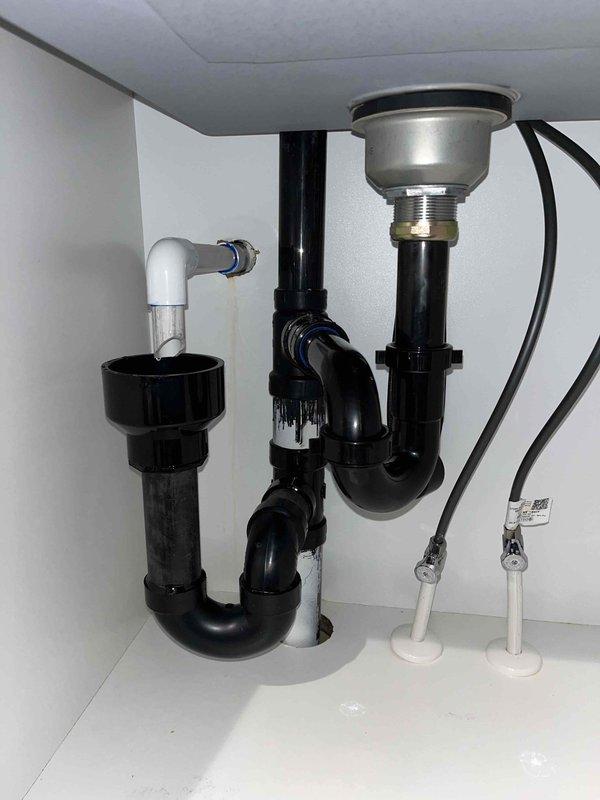 Responded to reported leak under sink in laundry room and inspected plumbing assembly beneath cabinet. Upon investigation, identified leak source at P-trap connections and observed water damage staining on interior cabinet wall. Evaluated black PVC drain assembly, chrome sink strainer, flexible supply lines, and all associated fittings to ensure proper configuration and seal integrity, with drip pan positioned to capture any residual moisture during assessment.