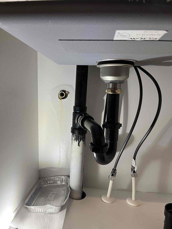 Responded to reported leak under sink in laundry room and inspected plumbing assembly beneath cabinet. Upon investigation, identified leak source at P-trap connections and observed water damage staining on interior cabinet wall. Evaluated black PVC drain assembly, chrome sink strainer, flexible supply lines, and all associated fittings to ensure proper configuration and seal integrity, with drip pan positioned to capture any residual moisture during assessment.