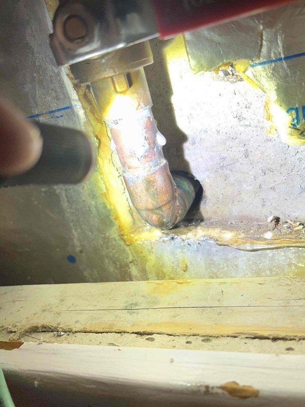 Responded to basement leak investigation. Upon inspection, identified heavily corroded copper pipe fitting beneath sink fixture with significant green oxidation and mineral deposits, causing active leak. Corroded connection showed advanced deterioration with failed yellow foam sealant around pipe penetration through cabinet base. Documented water damage to surrounding wooden surfaces and accumulation of moisture-related staining on adjacent plumbing components.
