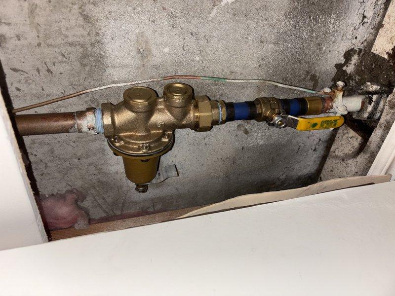 Responded to after-hours emergency call for leaking and broken water main shutoff valve in basement. Upon arrival, inspected 20+ year old plumbing system dating from 1989 mounted on front basement wall. Assessed brass valve assembly with multiple ports, copper supply line, blue-handled ball valve, and connecting brass compression fittings showing age-related wear and oxidation. Documented existing configuration including cylindrical brass valve body, pressure relief mechanism, and white PVC piping connections secured to concrete wall. System exhibited normal patina on copper components and signs of deterioration consistent with age of installation.