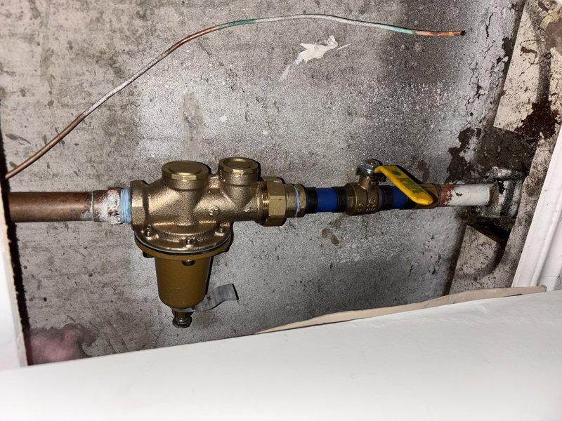 Responded to after-hours emergency call for leaking and broken water main shutoff valve in basement. Upon arrival, inspected 20+ year old plumbing system dating from 1989 mounted on front basement wall. Assessed brass valve assembly with multiple ports, copper supply line, blue-handled ball valve, and connecting brass compression fittings showing age-related wear and oxidation. Documented existing configuration including cylindrical brass valve body, pressure relief mechanism, and white PVC piping connections secured to concrete wall. System exhibited normal patina on copper components and signs of deterioration consistent with age of installation.