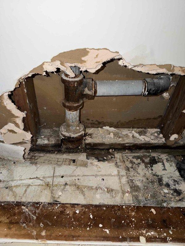 Responded to basement for initial sump pump installation consultation and assessment. Upon arrival, discovered extensive water damage throughout basement area including severely deteriorated walls, flooring, and exposed plumbing infrastructure. Documented significant moisture intrusion with visible mold growth along ceiling-wall junctions and dark staining patterns indicating long-term water exposure. Observed corroded galvanized metal drain pipes with heavy rust accumulation connected to gray PVC drainage line. Wall sections showed substantial drywall damage with exposed wooden framing and studs. Floor tiles exhibited heavy staining, discoloration, and debris accumulation with exposed subflooring visible in damaged areas. Assessed optimal sump pump basin location based on basement grade, existing drainage patterns, and proximity to discharge point. Recommended comprehensive moisture remediation and structural repairs prior to sump pump installation due to severity of existing water da