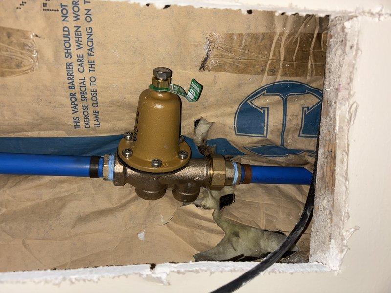 Responded to emergency call for leaking 2-year-old tank water heater in basement that caused partial flooding. Upon inspection, identified active leak originating from temperature and pressure relief (TPR) valve overflow pipe at top of unit. Observed corroded plumbing connections and compromised pressure regulation system with visible rust on metal fittings. Documented plumbing infrastructure including brass pressure regulator valve with blue water supply lines and deteriorated pipe connections showing significant corrosion. Captured photographic evidence of leak source and affected plumbing components for warranty and repair assessment purposes.