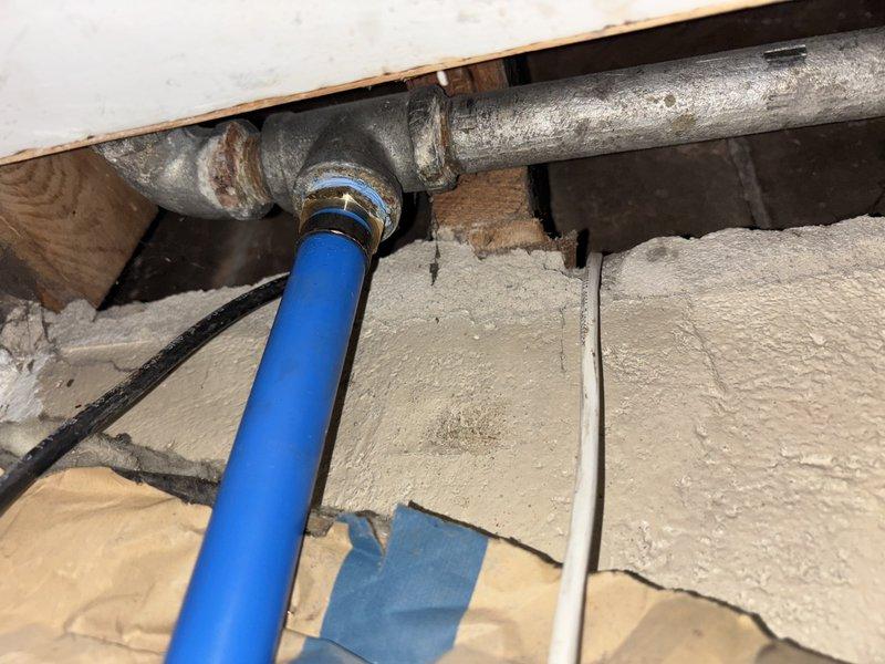 Responded to emergency call for leaking 2-year-old tank water heater in basement that caused partial flooding. Upon inspection, identified active leak originating from temperature and pressure relief (TPR) valve overflow pipe at top of unit. Observed corroded plumbing connections and compromised pressure regulation system with visible rust on metal fittings. Documented plumbing infrastructure including brass pressure regulator valve with blue water supply lines and deteriorated pipe connections showing significant corrosion. Captured photographic evidence of leak source and affected plumbing components for warranty and repair assessment purposes.