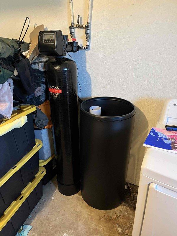 Completed installation of a 48,000 grain capacity water softener system with black twin-tank configuration. Mounted digital control head on primary treatment tank and connected white PVC bypass assembly with new isolation valves featuring color-coded handles for system control. Installed companion brine tank and filled with five bags of water softener salt for regeneration cycles. Programmed control head to customer's specific water hardness level for optimal softening performance. All plumbing connections secured and system tested for proper operation and cycle functionality.