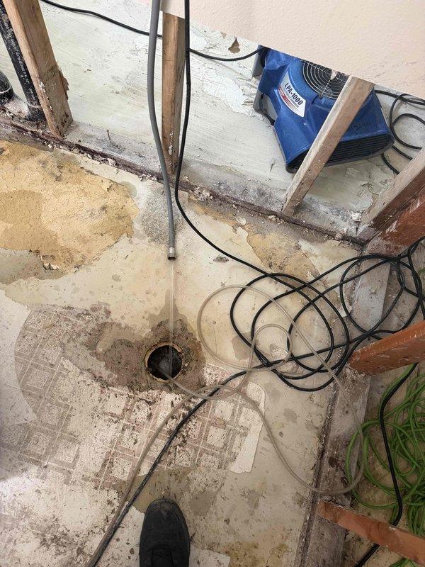 Responded to emergency drain cleaning request for basement bathroom floor drain experiencing backup issues. Upon arrival, found concrete floor with visible water staining and moisture damage, with commercial-grade air movers and drying equipment already in operation to address standing water conditions. Performed drain cleaning service on basement floor drain using cable auger equipment to clear blockage and restore proper drainage. Successfully cleared obstruction and verified drain flow was restored to normal operation.