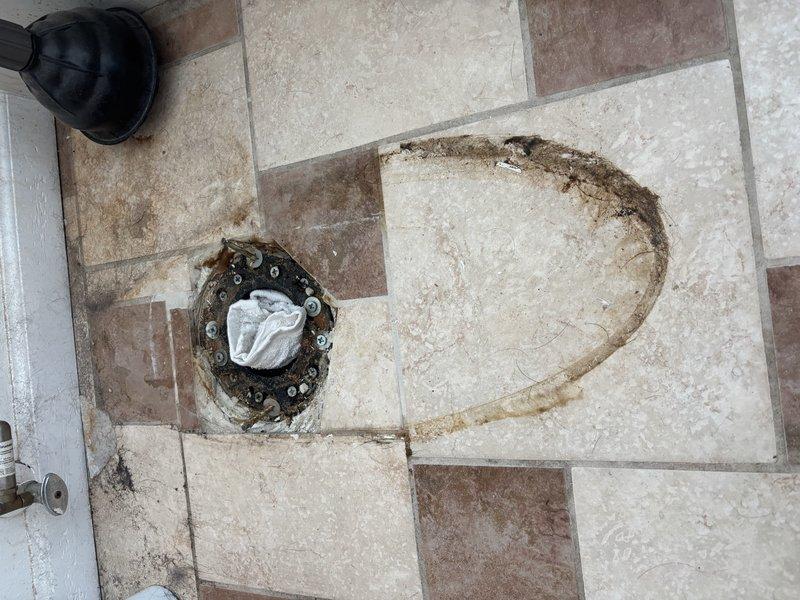 Responded to a known ceiling leak that was not resolved during a previous service visit. Upon arrival, accessed the bathroom area where another company had already opened the ceiling to expose the source of the water intrusion. Inspected the toilet flange assembly and surrounding floor area, noting significant water damage, staining, and deterioration around the toilet mounting location. Examined the exposed ceiling fixture above, which displayed extensive rust corrosion on the metal mounting plate and hardware, indicating prolonged moisture exposure. Identified compromised sealing at the toilet base as the primary leak source, with water migrating through the floor and into the ceiling below. Documented conditions with photographs showing the corroded ceiling-mounted components and water-damaged flooring. Customer also reported a potential secondary leak in the laundry room requiring further investigation.