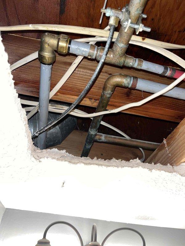 Responded to emergency call for burst bathroom pipe with active water seepage causing property damage. Upon arrival, confirmed main water supply had been shut off by occupant. Created ceiling access point to expose damaged plumbing infrastructure, revealing compromised pipe connection at brass elbow fitting where older brass/copper piping met PVC supply lines. Identified leak source at corroded brass fitting junction between floor joists. Assessed extent of water damage to ceiling drywall and surrounding structural members. Documented existing plumbing configuration showing aged brass components with visible patina and corrosion throughout the system. Secured exposed piping with plastic strapping to joists and verified water supply remained off to prevent further seepage while permanent repairs could be scheduled.