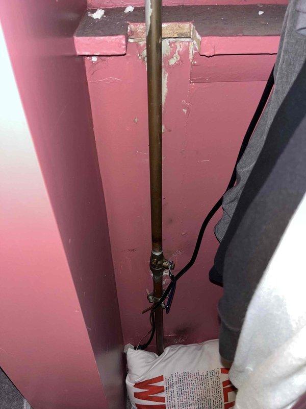 Responded to emergency call for burst bathroom pipe with active water seepage causing property damage. Upon arrival, confirmed main water supply had been shut off by occupant. Created ceiling access point to expose damaged plumbing infrastructure, revealing compromised pipe connection at brass elbow fitting where older brass/copper piping met PVC supply lines. Identified leak source at corroded brass fitting junction between floor joists. Assessed extent of water damage to ceiling drywall and surrounding structural members. Documented existing plumbing configuration showing aged brass components with visible patina and corrosion throughout the system. Secured exposed piping with plastic strapping to joists and verified water supply remained off to prevent further seepage while permanent repairs could be scheduled.