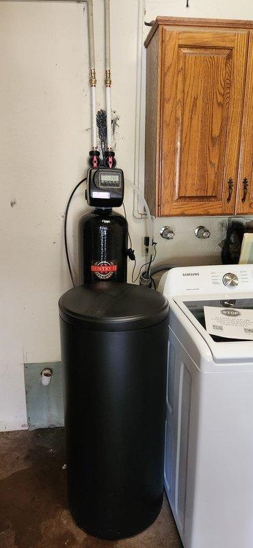 Installed 48,000 grain capacity water softener system with new brine tank in residential utility room. Connected unit to existing plumbing with white PEX supply lines and brass fittings, mounted black control valve with digital display, and filled brine tank with five bags of salt. Performed minor copper pipe repair during installation and programmed system settings to match house water hardness levels.